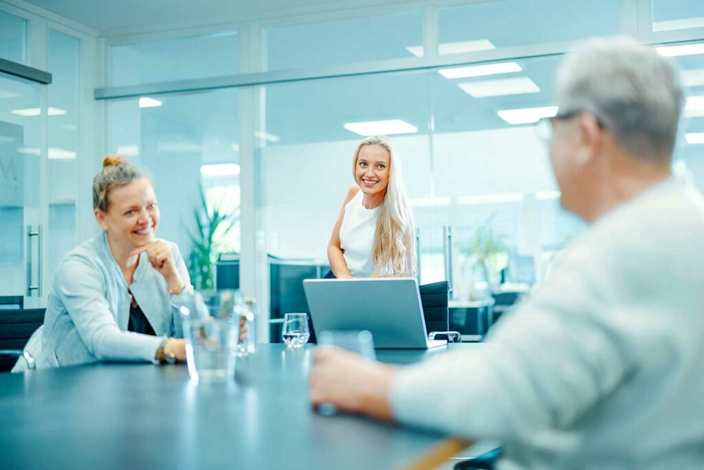 Drei Personen sitzen in einem modernen Besprechungsraum an einem Konferenztisch, lachen und unterhalten sich freundlich.