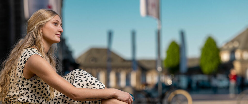 Frau mit langem blondem Haar sitzt im Sommerkleid im Stadtbereich und blickt zur Seite, im Hintergrund verschwommene Gebäude und Bäume.
