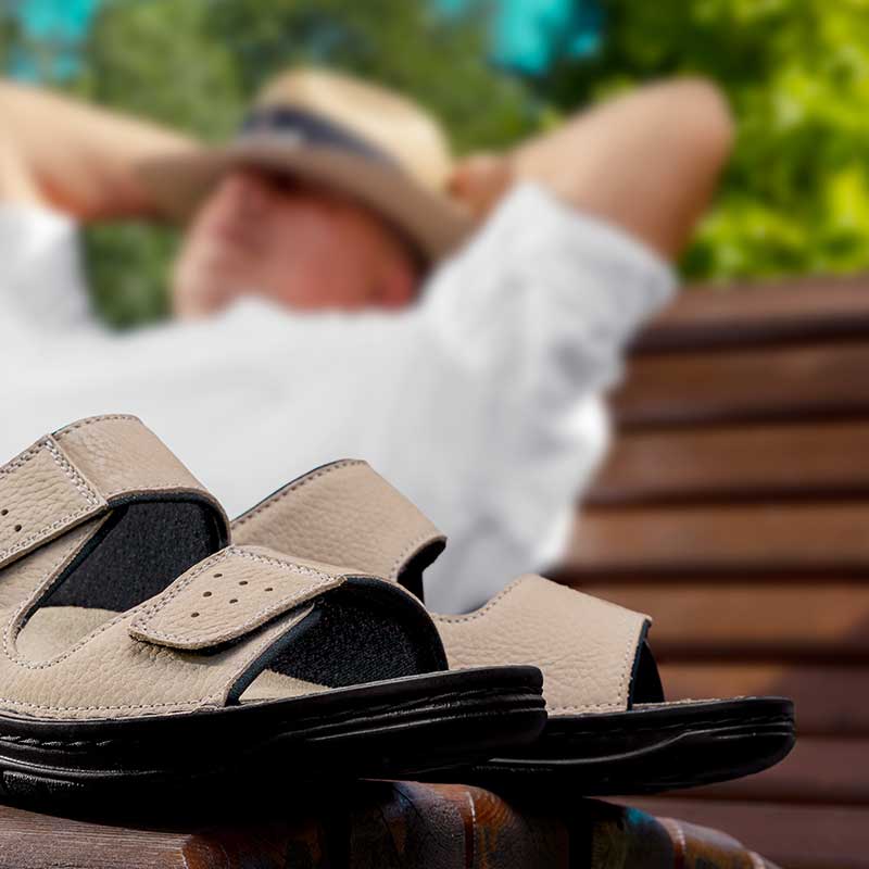 Beige Berkemann-Pantoletten Tobias mit schwarzer Sohle liegen auf einer Bank, im Hintergrund entspannt ein Mann mit Hut.