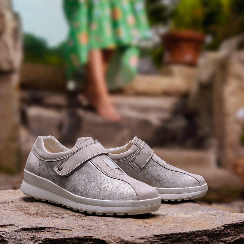 Silbergraue Berkemann-Schuhe Susanna mit Klettverschluss stehen auf einem Stein, im Hintergrund unscharf eine Person in grünem Kleid.