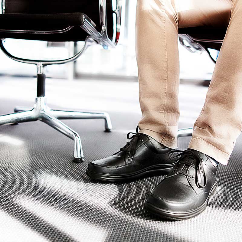 Schwarze Berkemann-Schnürschuhe an den Füßen einer sitzenden Person mit beigefarbener Hose, in einem modernen Büro mit Drehstuhl.