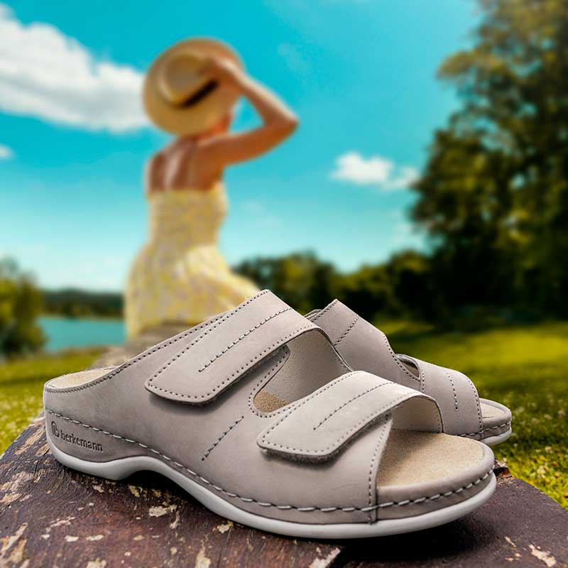 Beige Berkemann-Pantoletten Daria mit zwei Klettverschlüssen stehen auf einem Holzstamm, im Hintergrund sitzt unscharf eine Frau im gelben Kleid und Sonnenhut am See.