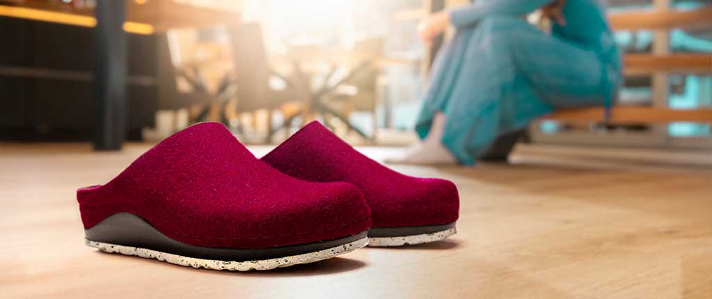 Rote Berkemann-Hausschuhe Blanche stehen auf einem Holzboden vor einer Treppe, im Hintergrund ist unscharf eine Person in einem blauen Kleid zu sehen.