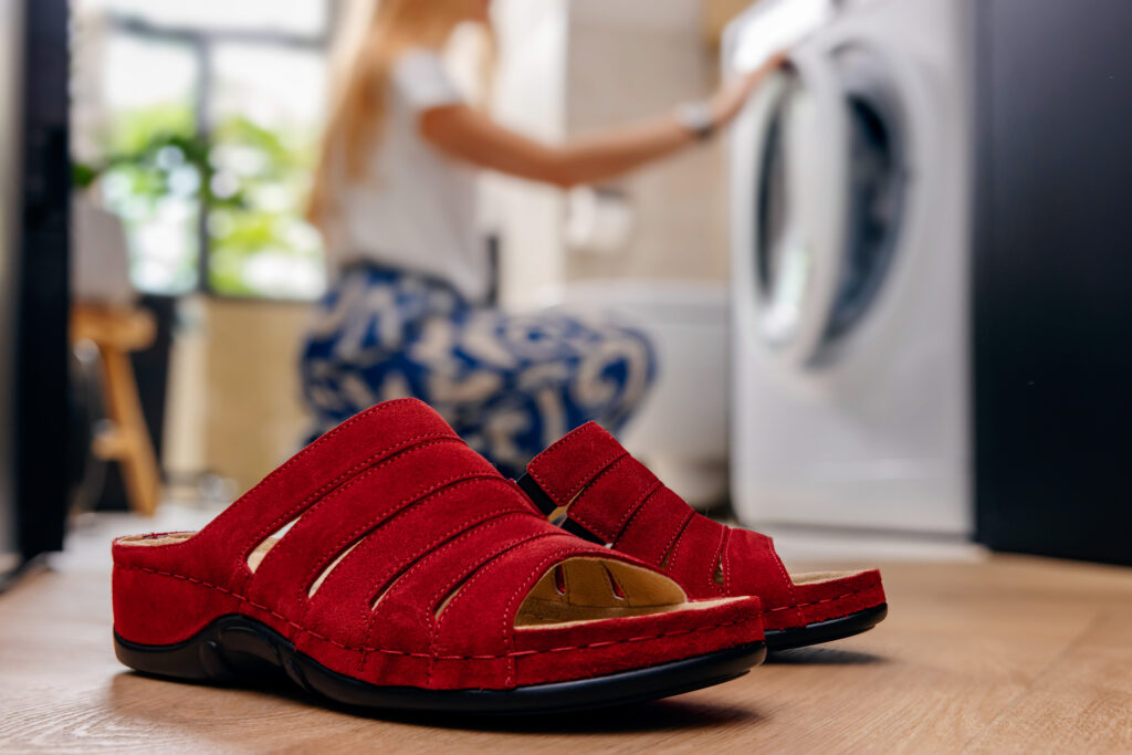 Berkemann-Pantolette Bine in Rot mit weichem Leder und bequemer Sohle. Im Hintergrund kniet eine Frau vor einer Waschmaschine – wohnliche Alltagsszene mit Fokus auf den komfortablen, pflegeleichten Schuh im Vordergrund.