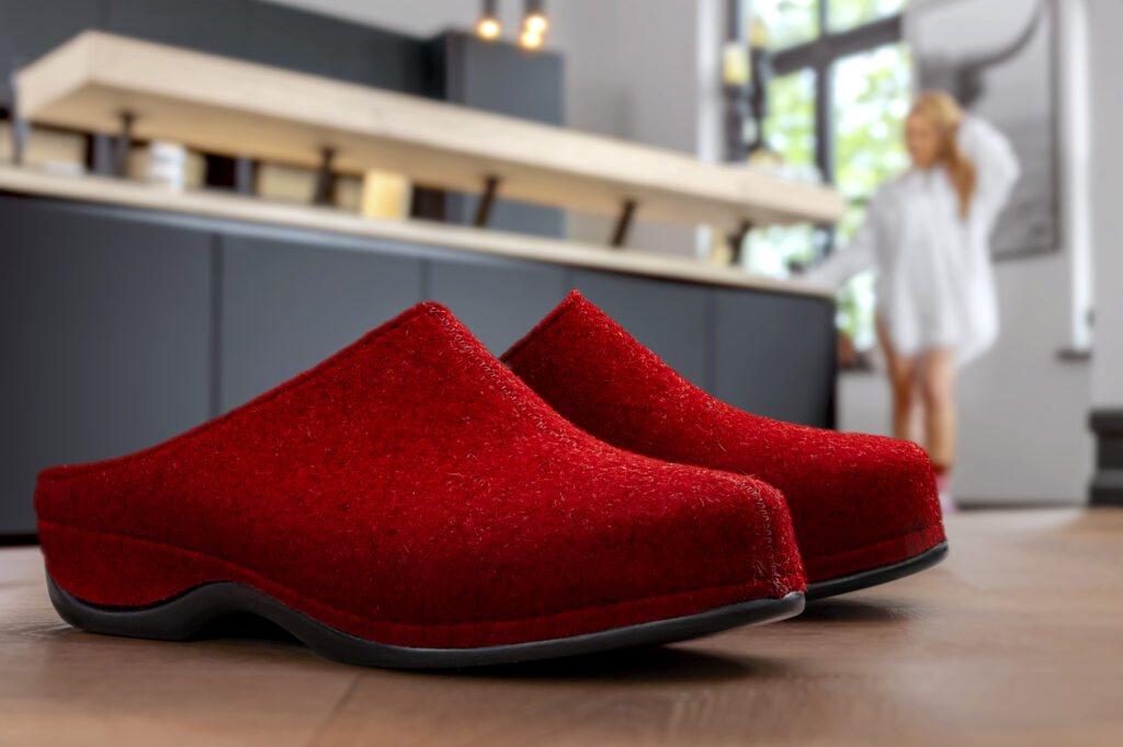 Berkemann-Hausschuh Florina in Rot mit ergonomischer Laufsohle. Im Hintergrund steht eine Frau in heller Kleidung in einer modernen Küche – gemütliche Alltagsszene mit Fokus auf den warmen, komfortablen Filzschuh im Vordergrund.