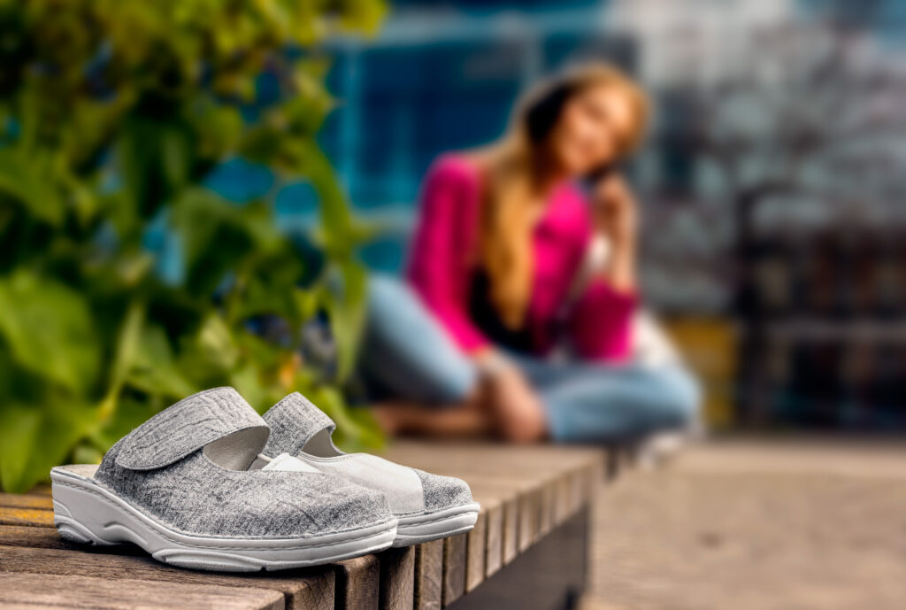 Graue Berkemann-Pantoletten, Modell Heliane, mit breitem Klettverschluss und heller Sohle, stehend auf einer Holzbank im Freien; im Hintergrund sitzt eine Person in Jeans und pinkfarbener Jacke entspannt in der Sonne.