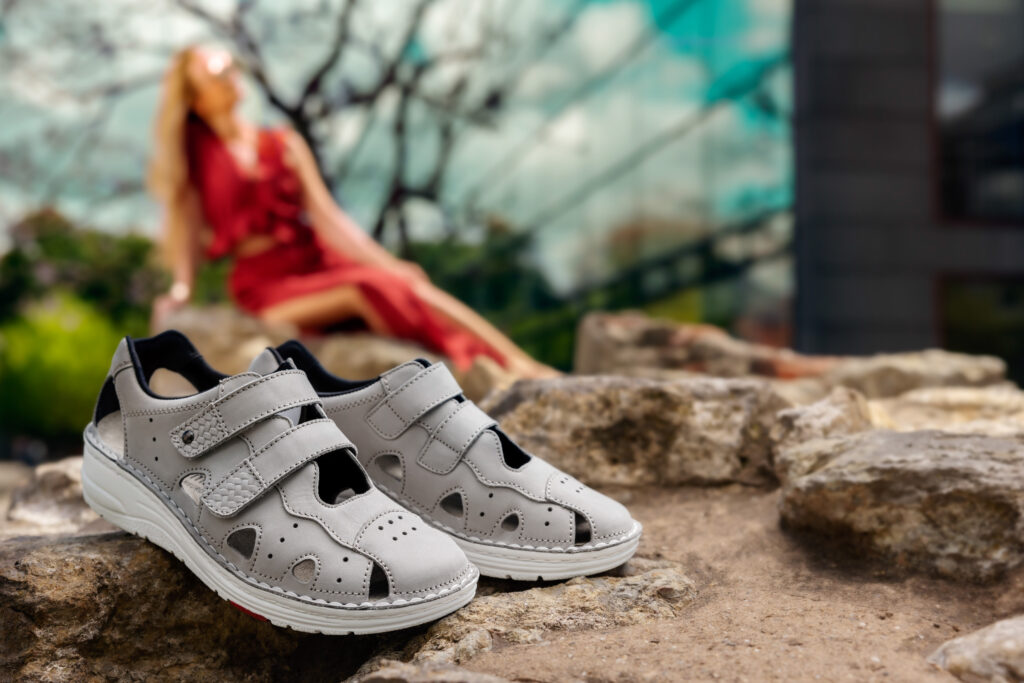 Graue Berkemann-Sandalen, Modell Larena, mit zwei Klettverschlüssen und perforierten Details, stehend auf Felsen im Freien; im Hintergrund sitzt eine Person in einem roten Kleid entspannt in der Sonne.