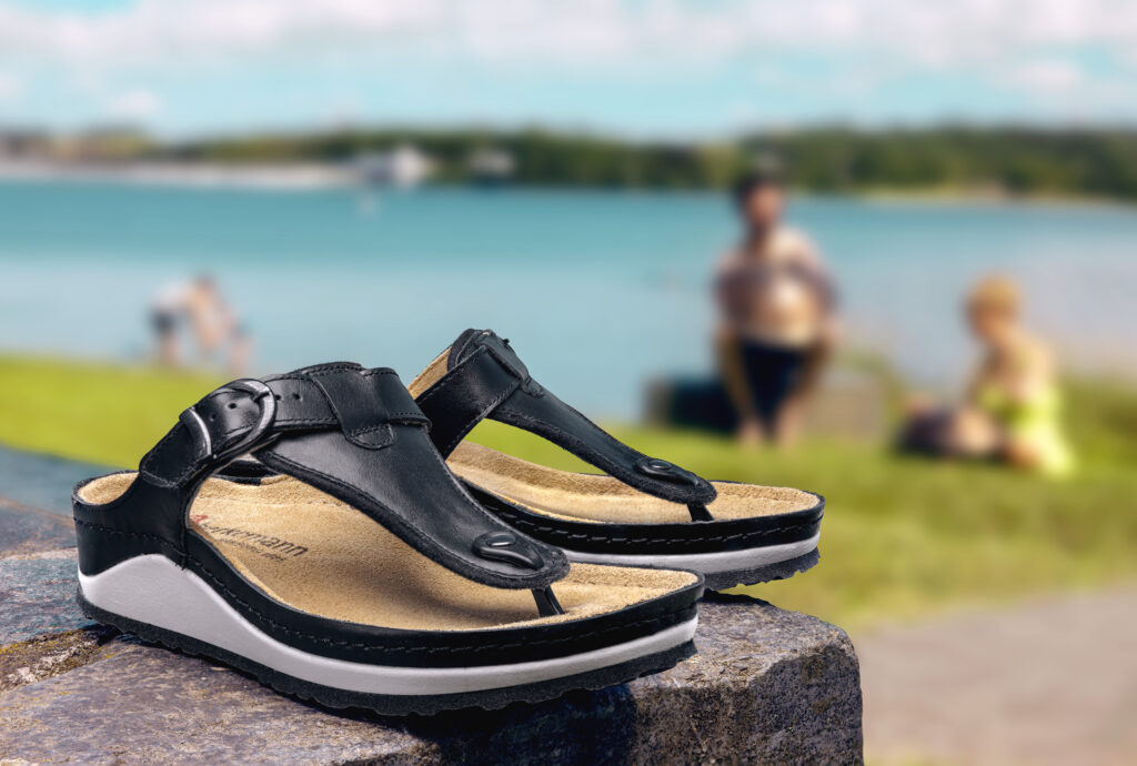 Schwarze Berkemann-Zehenstegsandalen, Modell Mila, mit verstellbarem Riemen und heller Sohle, stehend auf einem Stein am Seeufer; im Hintergrund entspannen Menschen am Wasser bei sonnigem Wetter.