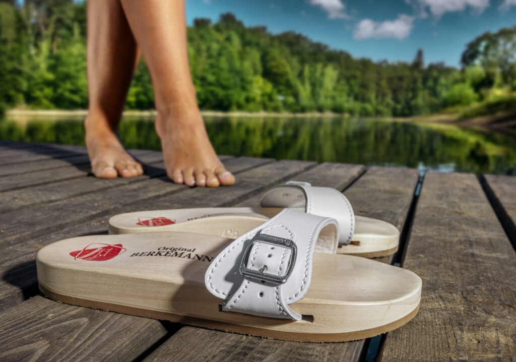 Ein Paar weiße Berkemann-Originalsandalen mit Holzfußbett liegt auf einem hölzernen Steg an einem See. Im Hintergrund steht eine Person barfuß, umgeben von grüner Natur und blauem Himmel mit Wolken.