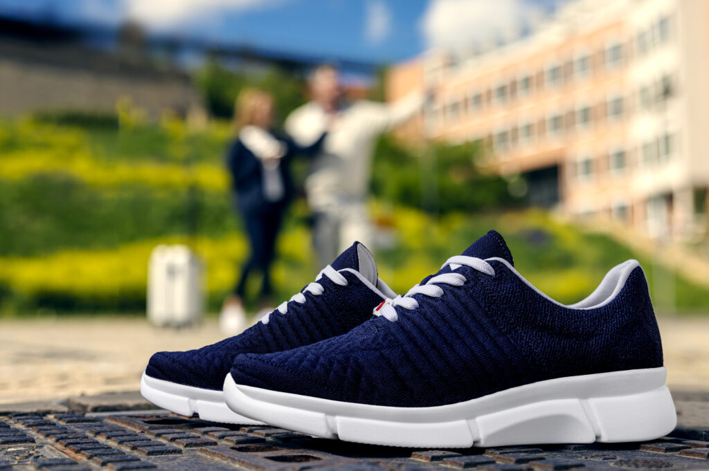 Dunkelblaue Berkemann-Sneaker, Modell Pinar, aus atmungsaktivem Strickmaterial mit weißer Sohle, stehend im Freien auf einem Gitterrost; im Hintergrund unterhalten sich zwei Personen mit Gepäck vor einem modernen Gebäude.