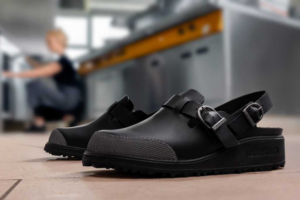 Berkemann-Arbeitsclog X-Pro Maxor in Schwarz aus der Work-Linie, mit robuster Laufsohle, Zehenschutzkappe und verstellbarem Fersenriemen. Im unscharfen Hintergrund arbeitet eine Person in einer professionellen Küche.