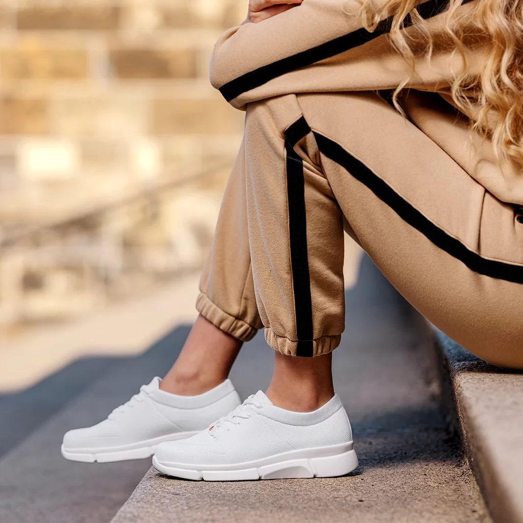 Weiße Berkemann-Sneaker Allexis an den Füßen einer Frau, die in beigefarbenen Jogginghose auf einer Treppe sitzt.