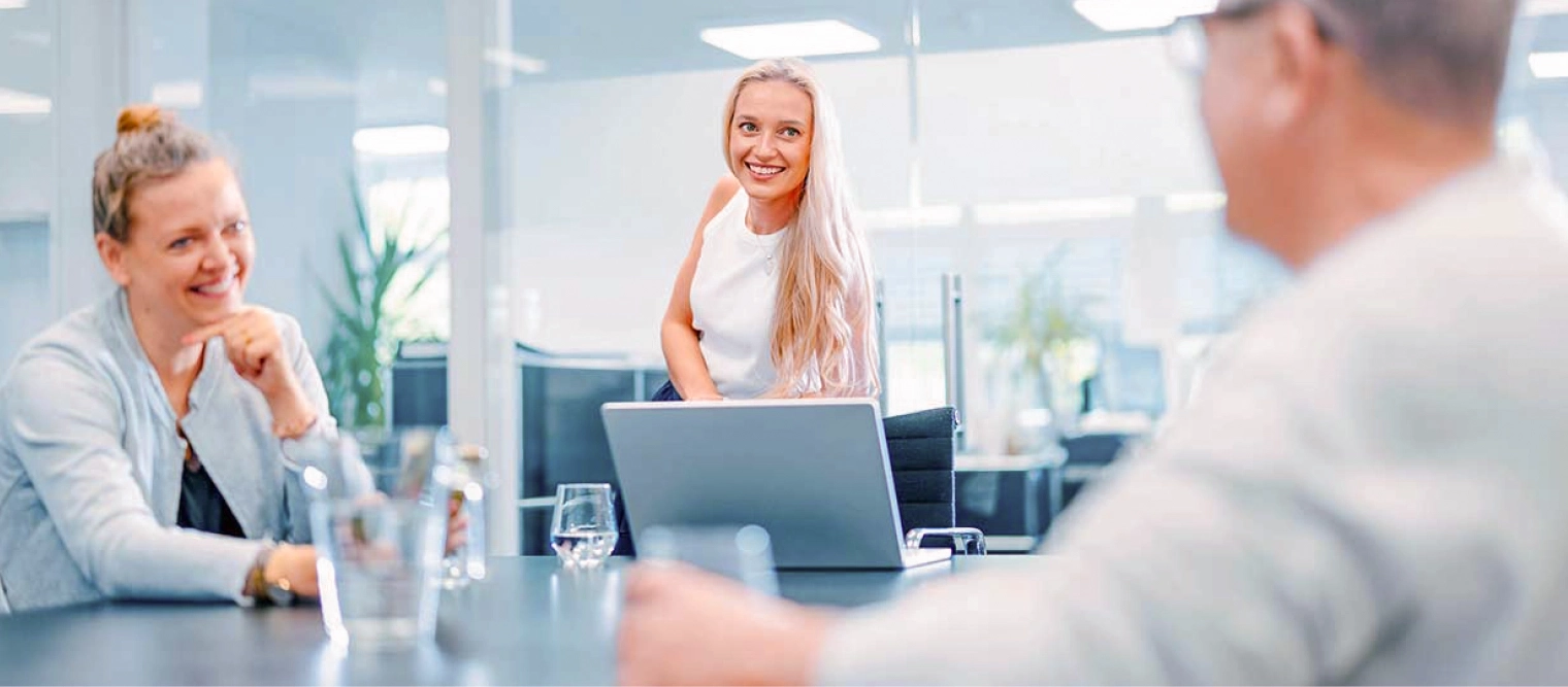 Drei Personen sitzen in einem modernen Besprechungsraum an einem Konferenztisch, lachen und unterhalten sich freundlich.
