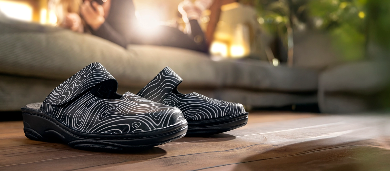 Schwarze Clogs von Berkemann, Modell Heliane, mit weißem Linienmuster auf Holzboden; im Hintergrund unscharf eine Frau, die auf dem Sofa liegt.