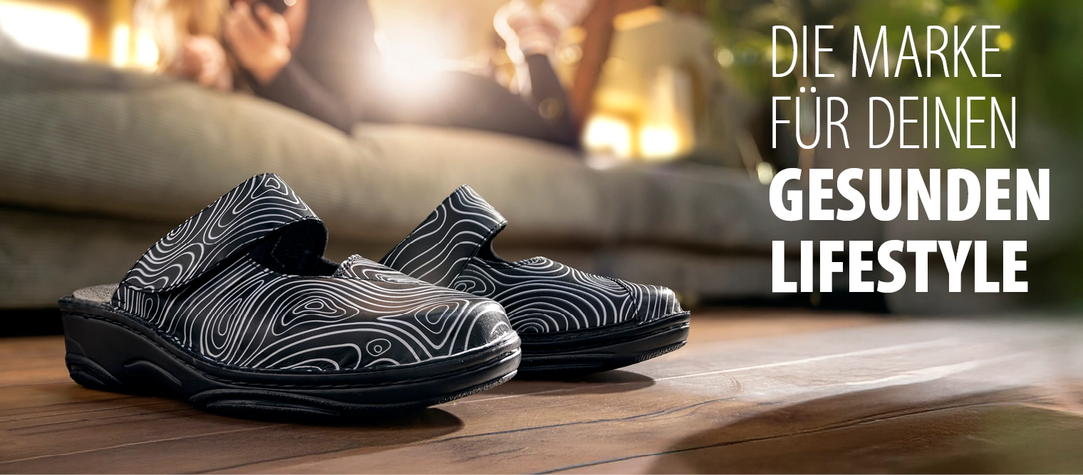 Schwarze Clogs von Berkemann, Modell Heliane, mit weißem Linienmuster auf Holzboden; im Hintergrund unscharf eine Frau, die auf dem Sofa liegt.