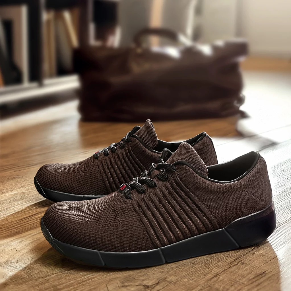 Brauner Berkemann-Sneaker Björn aus Strickmaterial mit schwarzer Sohle, stehend auf einem Holzboden, im Hintergrund unscharf eine braune Tasche.