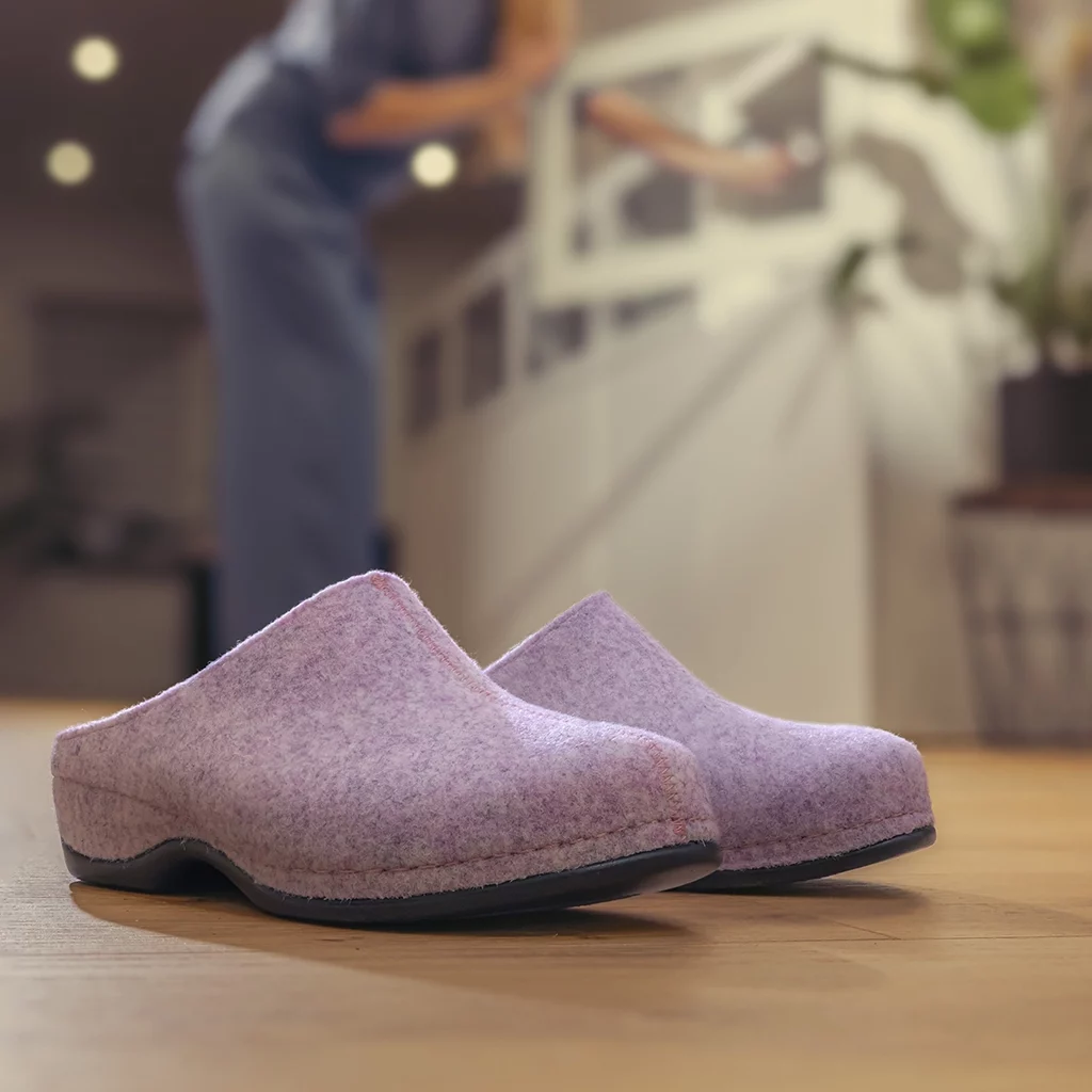 Rosafarbene Filzhausschuhe von Berkemann, Modell Florina, mit schwarzer Sohle, stehend auf einem Holzfußboden; im Hintergrund eine Person in blauer Kleidung, die sich an einem Geländer abstützt.