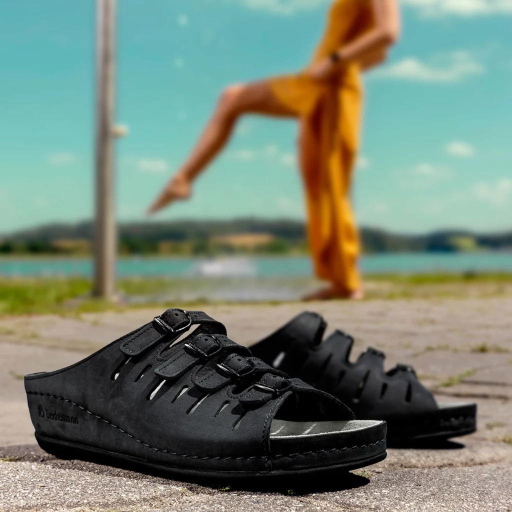 Berkemann-Pantolette Hassel mit vier verstellbaren schwarzen Lederriemen und offener Spitze in Nahaufnahme, im Vordergrund auf Pflasterstein, unscharfe Person im gelben Jumpsuit im Hintergrund.