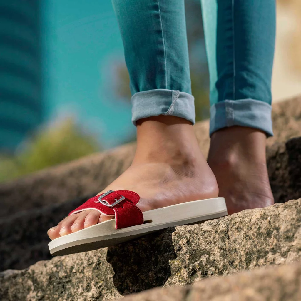 Rote Berkemann Original-Sandale mit Holzsohle an einem Fuß auf einer Steintreppe, Person trägt hochgekrempelte Jeans.
