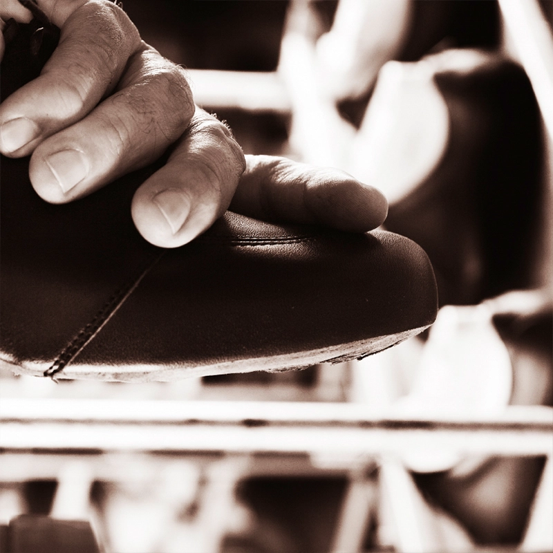 Nahaufnahme einer Hand, die bei der handwerklichen Schuhherstellung ein Lederschuh-Oberteil formt; Werkstattumgebung unscharf im Hintergrund.