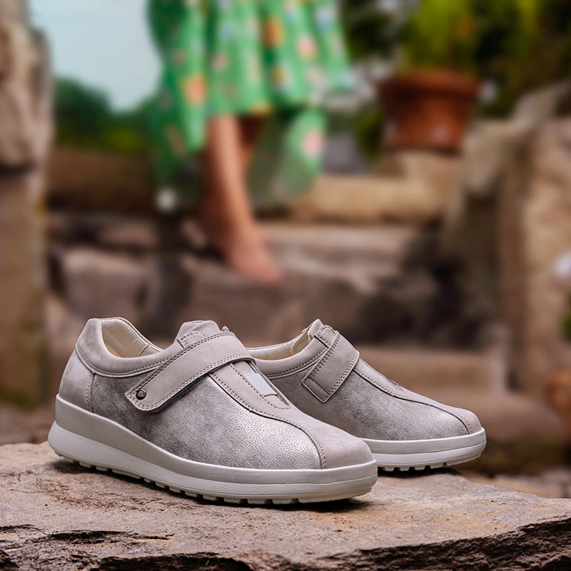 Berkemann-Schuh Susanna in Hellgrau mit breitem Klettverschluss, seitlich auf Stein platziert, unscharfe Person im grünen Kleid im Hintergrund.