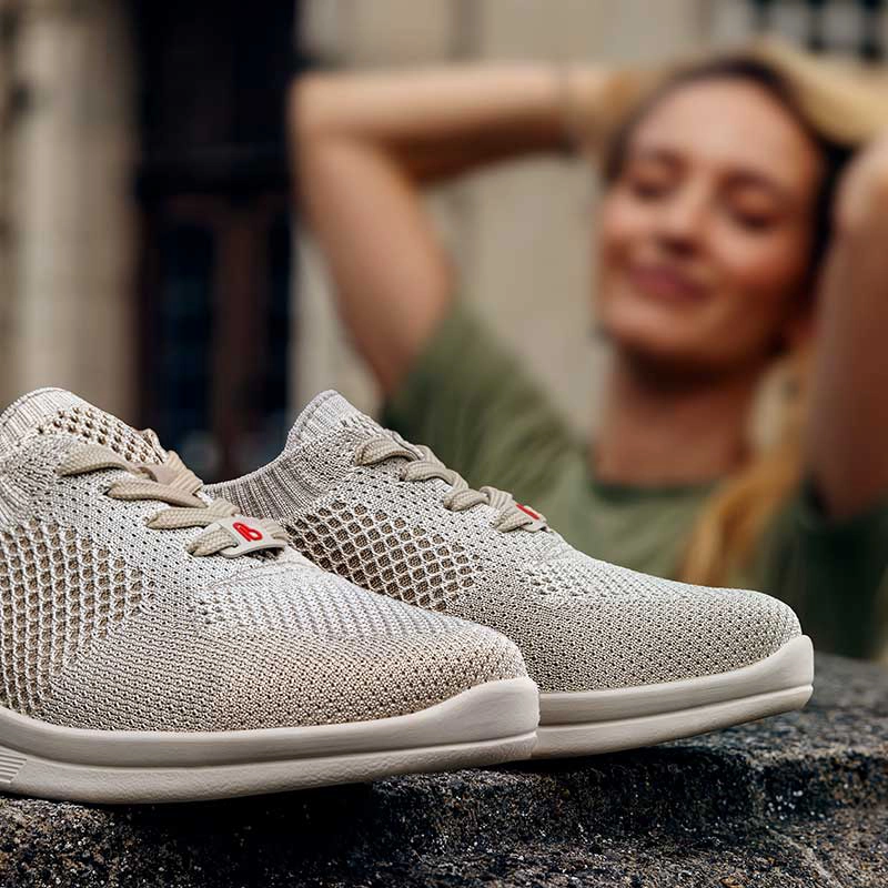 Beiger Strick-Sneaker von Berkemann, Modell Elea, in Nahaufnahme auf Steinfläche; im Hintergrund unscharf eine Frau in grüner Kleidung – sommerliche Alltagsszene im Freien.