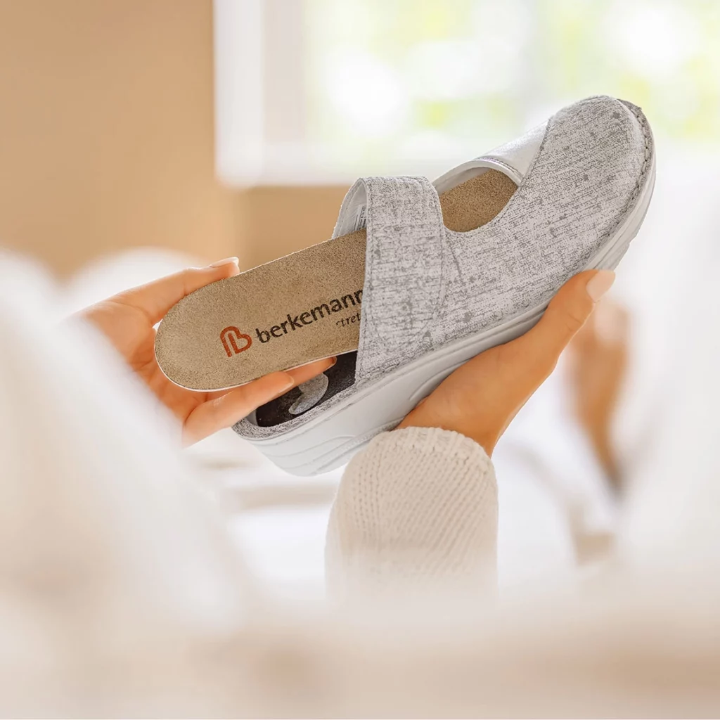 Person hält die graue Berkemann-Pantolette Heliane mit herausnehmbarem Wechselfußbett in den Händen, Nahaufnahme in heller Wohnumgebung.