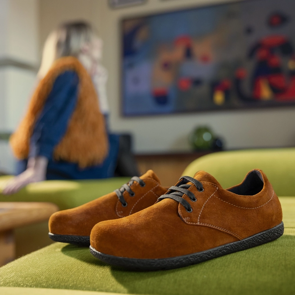 Berkemann Schnürschuh Karlina aus orangefarbenem Veloursleder in Seitenansicht auf grünem Polster, schwarze Laufsohle; im Hintergrund eine Person im Wohnraum.
