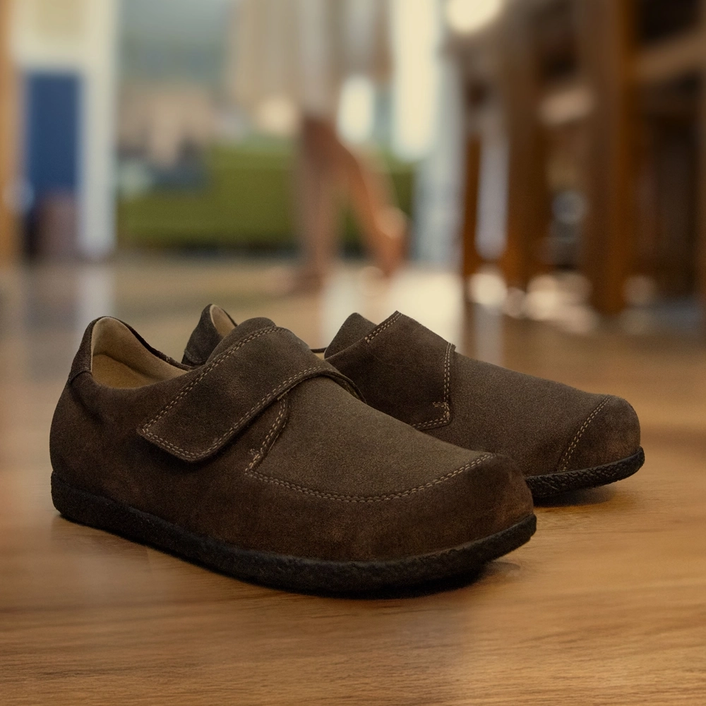 Halbschuh Renea von Berkemann aus braunem Veloursleder und Stretch mit Klettverschluss steht im Vordergrund; im Hintergrund eine Person im Wohnraum.