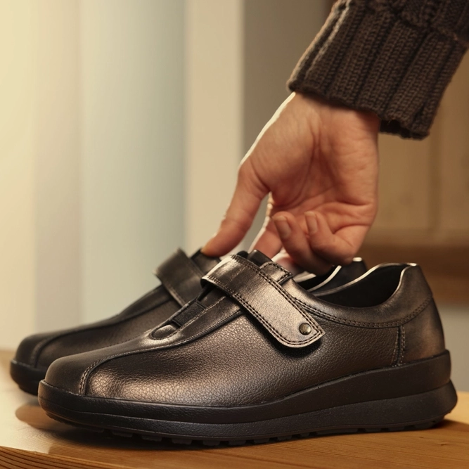 Berkemann Halbschuh Susanna aus bronzebraunem Leder mit Klettverschluss in Seitenansicht auf Holzstufe; Hand greift nach dem Schuh, Treppenbereich im Hintergrund.