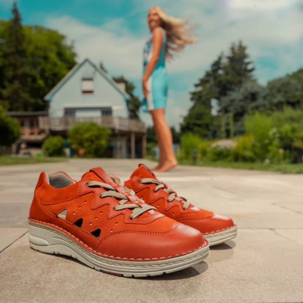 Orangefarbene Schnürschuhe Laurie von Berkemann stehen auf einer Terrasse vor einem Haus; im Hintergrund läuft eine barfüßige Person vorbei.