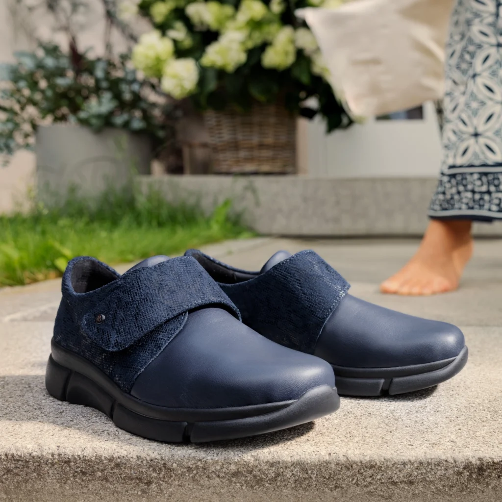 Zwei dunkelblaue Halbschuhe Sanela von Berkemann mit Stretch und Klettverschluss stehen auf einer Stufe vor einem Hauseingang; im Hintergrund ist eine barfuß stehende Person zu sehen.