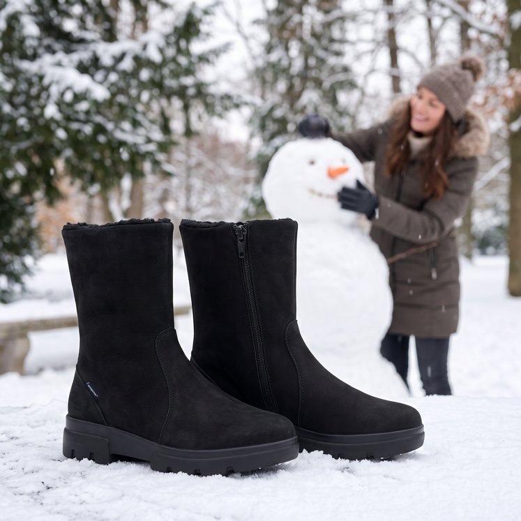 Schwarze Winterstiefel mit Sympatex im Schnee, im Hintergrund baut eine Frau in winterlicher Waldkulisse einen Schneemann.