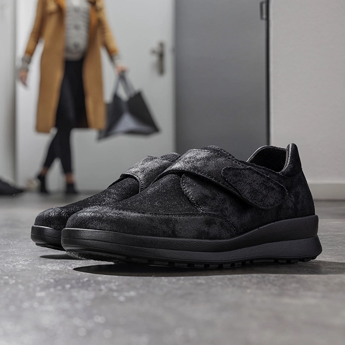 Schwarze Berkemann-Schuhe Modell Venita stehen im Vordergrund; im Hintergrund betritt eine Person den Flur mit Einkaufstaschen.