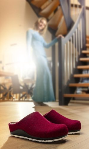 Rote Berkemann-Hausschuhe Blanche stehen auf einem Holzboden vor einer Treppe, im Hintergrund ist unscharf eine Person in einem blauen Kleid zu sehen.